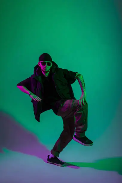 Photo of Stylish trendy professional man dancer with sunglasses and hat in fashion black clothes dancing vogue style in creative color studio with green and pink light