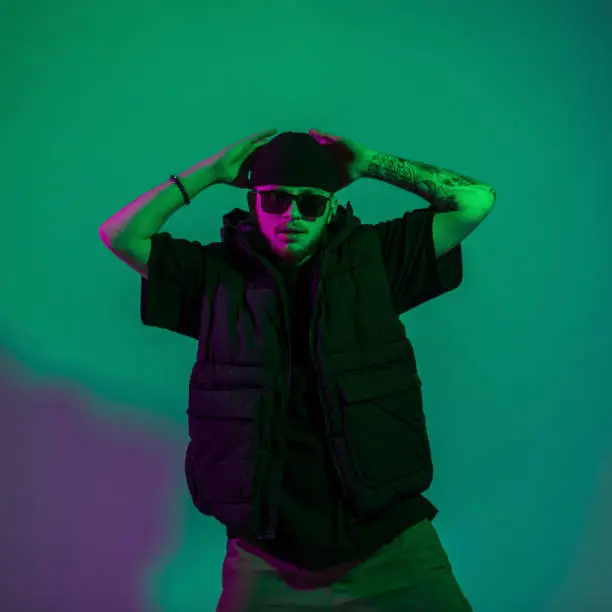 Photo of Cool fashionable young hipster man with vintage sunglasses in trendy stylish outfits poses in creative colored studio with green and pink light