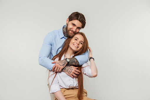 Happy fashionable beautiful couple in the studio. Stylish hipster handsome man with beard and tattoo hugging happiness redhead beautiful girl with smile on white background