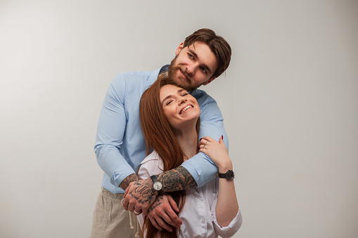 Beautiful fashionable happy couple in the studio. Fashion handsome brutal man with smile, tattoos and beard hugging beautiful stylish redhead woman in shirt on white background