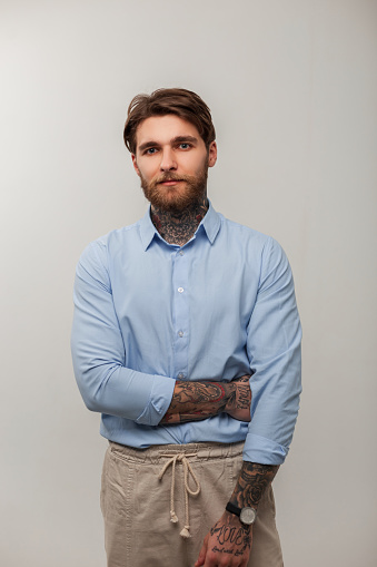 Hipster brutal handsome man with beard, hairstyle and tattoos in fashion blue shirt stands in a gray background