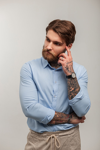 Business handsome brutal hipster man with a beard and a tattoo in a fashion blue shirt is standing and thinking against a gray background in the studio