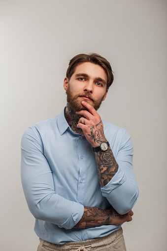Stylish successful man hipster businessman with beard and tattoos in blue fashion shirt and thinking in studio on white background