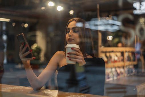 Beautiful young woman in a fashion black top sits near a window in a cafe, drinks coffee and watches the news feed on a smartphone.