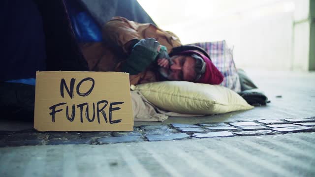 Homeless beggar man lying outdoors in city, no future cardboard sign near him.