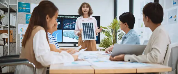 Photo of Asian business people team brainstorm meeting in sustainable corporate office. Presentation ESG project planning, solar panel, wind turbine power energy generation. Environmental conservation concept