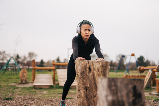 adult Asian woman is training in the gym outdoors, she is wearing headphones and leaning on a tree stump.