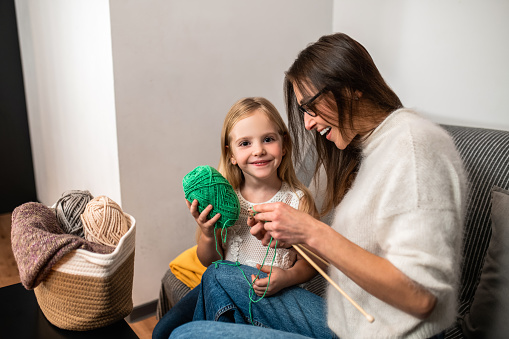 Young mother teaching her daughter to knot on sofa in home interior knitting with her daughter