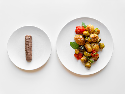 Two white plates on table, one with single chocolate bar and other with roasted baby potatoes and Brussels sprouts, contrasting healthy and indulgent food choices. High quality photo