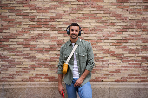 Portrait of millennial stylish man looking happy at camera with wireless headphones holding phone leans against brick wall. Handsome male person posing with confident attitude outdoors. Copy space