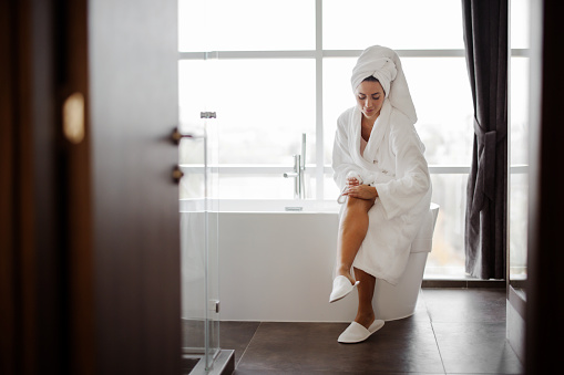 Woman In Bathrobe Moisturizing Smooth Legs With Moisturizer Cream Sitting On Bathtub In Modern Bathroom Indoors After Morning Shower. Beauty, Skincare And Pampering Concept