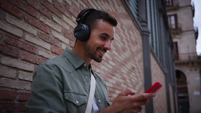 Side attractive young Caucasian man with headphones using cell phone leaning against brick wall.