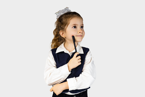 A happy little girl in a school uniform is standing with a smile, holding a pen in her hand. Her dress shirt has a collar and sleeves, and she is making a gesture with her thumb