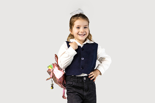 The little girl, with a smile on her face, is wearing a school uniform with puffed sleeves and carrying a backpack. She looks happy and her thumbs are hooked around her waist, making a cute gesture