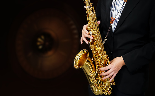 Male Musician in a Formal Black Suit Holds a Tenor Saxophone on blurred speaker background.