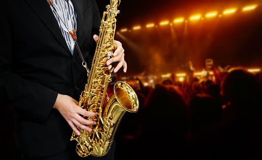 Male Musician in a Formal Black Suit Holds a Tenor Saxophone on the background of the concert atmosphere. Saxophonist Plays Jazz. Saxophone Close-up. Copy space