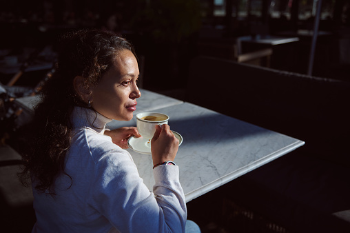 Confident and attractive curly haired young woman looking aside while sitting at coffee shop, enjoying her coffee break. People. Food and drink consumerism concept
