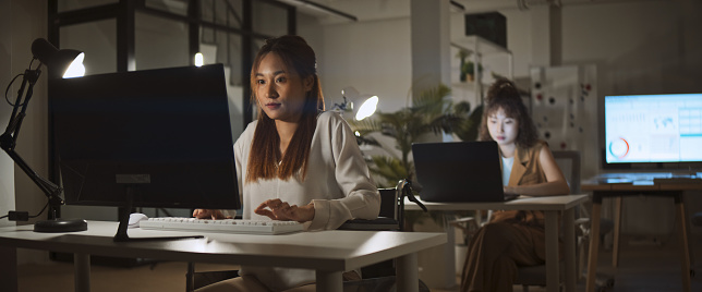 Young adult Asian businesswoman work late together with coworker at night in sustainable office, using desktop and laptop computer. Corporate business, overworked millennial generation z woman concept
