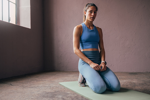 A pensive young woman in athletic wear sits on a yoga mat, resting after a workout in a calm studio environment.