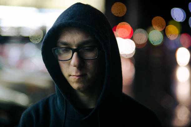 A young male caucasian person standings at nighttime in an urban city. He is wearing a black hood and looks depressed.
