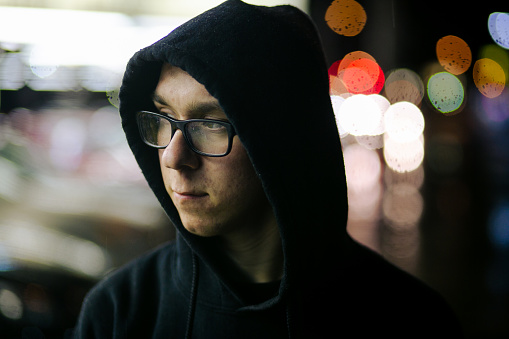 A young male caucasian person standings at nighttime in an urban city. He is wearing a black hood and looks depressed.