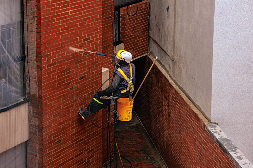 Bogotá, Colombia - February 19, 2024: A construction worker, is seen abseiling down a residential building to clean, repair and handle maintenance work. He is seen seated on his suspended seat. He is wearing waterproof clothing, rubber gloves and wellington shoes to prevent being drenched by the water he is spraying onto the brick wall.