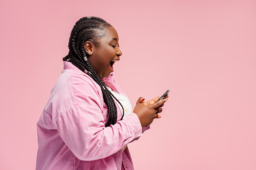 Happy African woman holding smartphone using mobile app, shopping online with sale, cash back isolated on pink background, copy space. Overjoyed female sports betting, win money, reading good news