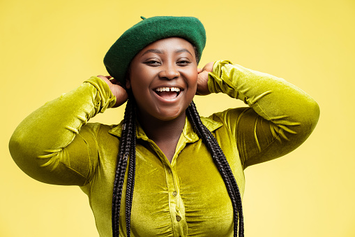 Beautiful smiling plus size fashion model with stylish braids hair, wearing green beret isolated on yellow background. Portrait happy African woman posing in trendy outfit in studio. Shopping, sale