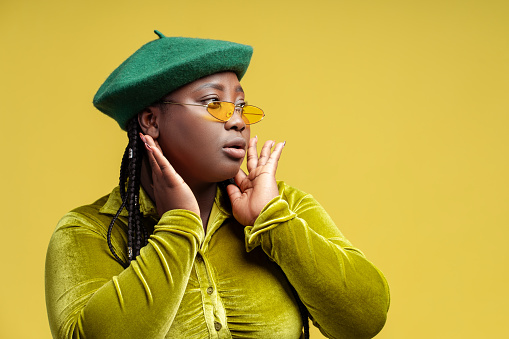 Beautiful modern African American woman wearing stylish casual clothes green beret, stylish eyeglasses looking away. Cute, cheerful fashion model posing isolated on yellow background. Shopping concept