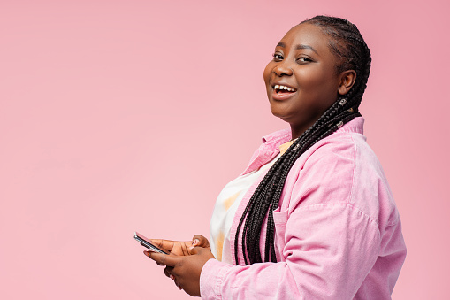 Smiling plus size African American woman holding mobile phone using mobile app looking at camera isolated on pink background, copy space. Attractive businesswoman ordering. Concept of online shopping