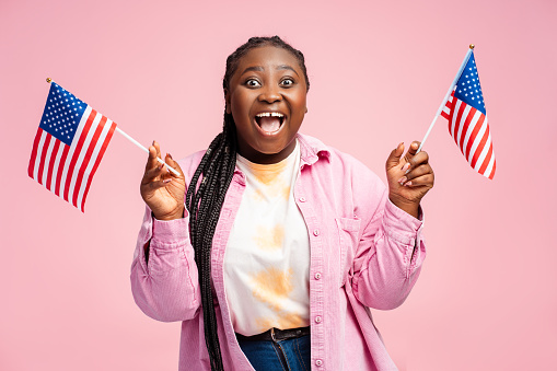Beautiful, overjoyed African American woman wearing casual clothes holding American flags looking at camera. Happy body positive supporter isolated on pink background. Election day concept