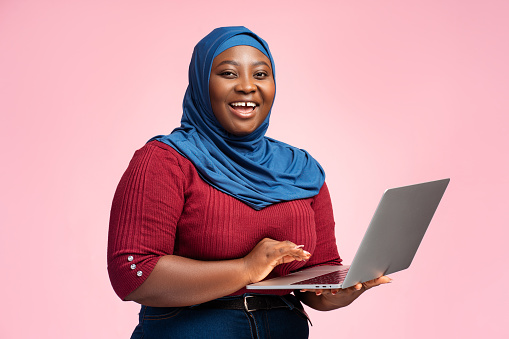 Beautiful, smiling Arab woman wearing traditional hijab holding laptop, ordering, looking at camera, standing isolated on pink background. Online technology concept