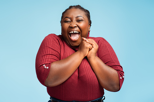 Beautiful, overjoyed African American woman rejoicing with open mouth looking at camera standing isolated on blue background. Concept of celebration, success