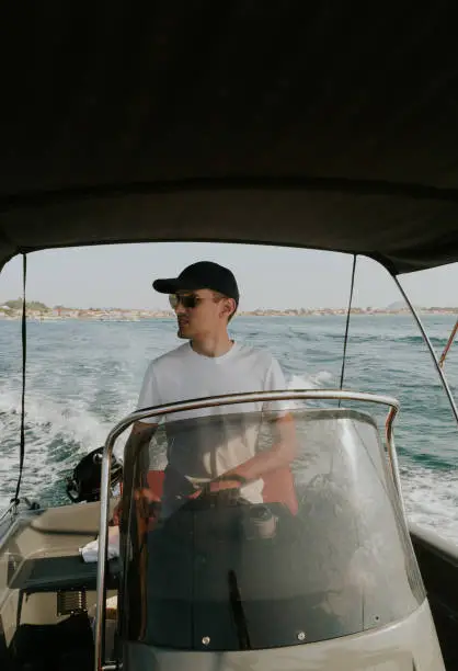 One young Caucasian guy in a cap and sunglasses sails alone on the sea on a boat, steering the helm and looking to the left, on a sunny summer day, close-up side view.