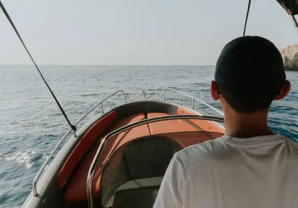 One young Caucasian guy in a cap and a white T-shirt stands from the back at the helm and sails alone on the sea on a boat, on a sunny summer day, close-up side view.