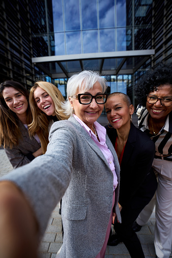 Vertical group of diverse business formal women taking selfie with phone looking smiling at frontal camera. Happy cheerful photo of adult female people co-workers outdoor. Gray mature lady in center