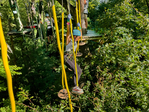 A people walks a rope bridge between trees in an amusement park in safety gear and a helmet. Rope park in wood forest.A group of people goes on hinged trail in extreme rope Park in summer forest.High-altitude climbing training on adventure track, equipped with safety straps.