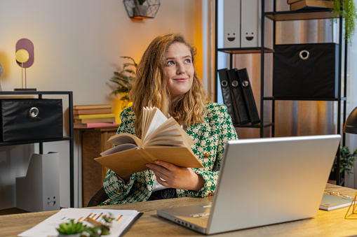 Caucasian young businesswoman reading interesting book, turning pages smiling enjoying literature, taking a rest after work. Happy cheerful freelancer girl with laptop relaxing at home office desk