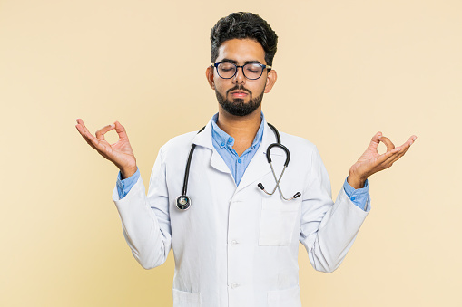 Keep calm down, relax, rest. Concentrated happy Indian doctor cardiologist man meditating breathes deeply, eyes closed peaceful mind taking a break. Arabian apothecary guy isolated on beige background
