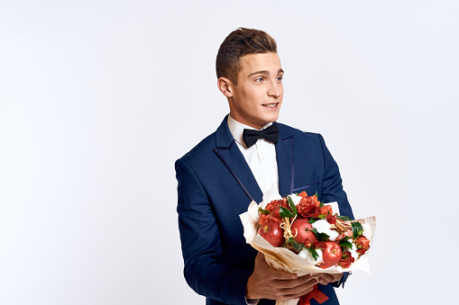 An elegant man in a classic suit with a bow tie holds a bouquet of flowers in his hand on a light background