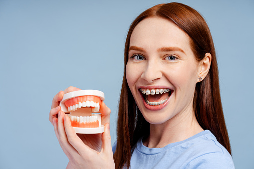 Closeup shot of a cheerful redhead woman with braces, holding a jaw explanatory model, isolated on a blue background. Emphasizes dental health concept