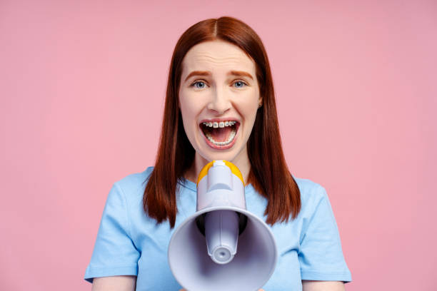 istock Ginger haired woman with braces, screaming through a megaphone, directly looking into the camera 2054160836