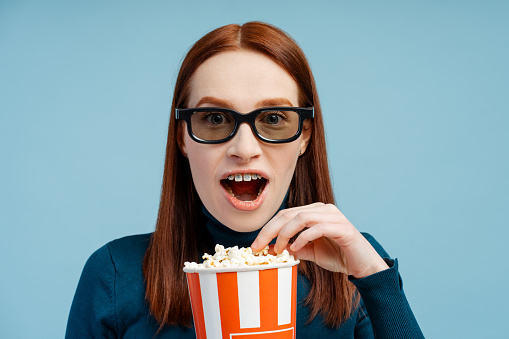 Portrait of an astonished ginger woman with braces, donning a polo neck sweater and 3D glasses, enjoying popcorn, mouth wide open, isolated on blue background