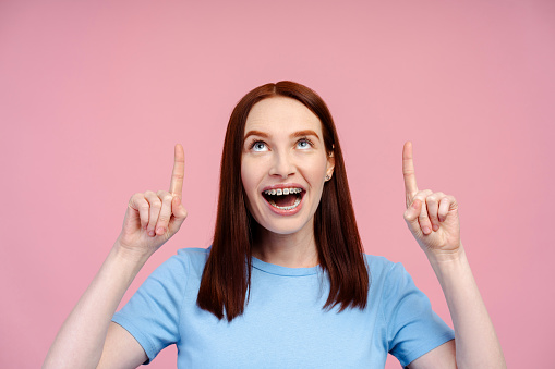 Cheerful, ginger haired woman wearing braces, in a T shirt, pointing up, isolated on a pink background. Ideal for marketing