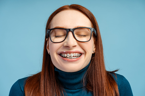 Picture of a stunning ginger haired woman with brackets and spectacles, clad in a polo neck jumper, standing with her eyes closed and a joyful smile, relishing the moment, isolated on blue background