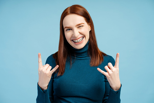 Funny overjoyed woman with braces wearing blue turtleneck showing rock sign looking at camera isolated on blue background. Positive lifestyle, emotions concept