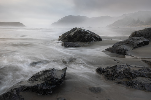 A rocky shoreline with a large rock in the foreground. The water is choppy and the sky is overcast, creating a moody and dramatic atmosphere. The rocks and water seem to be in constant motion