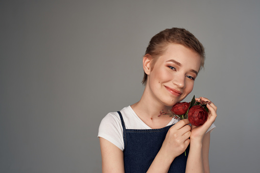 Portrait of charming young florist standing in the flower shop doorway and looking at beautiful red roses she is holding.