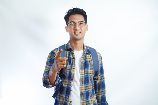 Happy fun young Asian man student in casual clothes bag glasses pointing finger at camera on you motivating encourage isolated on white background. high school university college concept