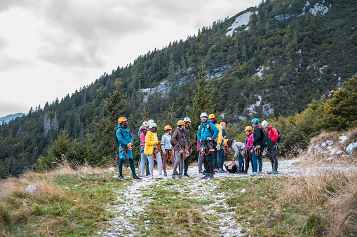 A diverse group of enthusiastic male and female hikers wearing helmets and harnesses prepares for a zipline adventure in a mountainous outdoor setting. The scene captures the group in casual outdoor attire against a backdrop of forested slopes and a hint of snow underfoot, highlighting the blend of excitement and adventure.